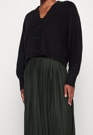 Femme portant un cardigan court en tricot noir boutonné avec un col en V et une jupe plissée taille haute vert foncé.
