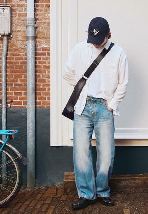 Muškarac u bijeloj majici, svijetloplavim trapericama, crnoj kapici i crnoj torbi preko ramena stoji na popločanom pločniku blizu bicikla i ciglene zidne površine.