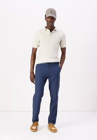 Creme kabelstickad kortärmad polo, marinblå lätta byxor och bruna sneakers med vita accenter, toppat med en grå keps.