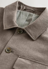 Chaqueta marrón con textura, cuello de solapa abierto, dos botones y un solo bolsillo en el pecho. Interior forrado con tela gris clara.