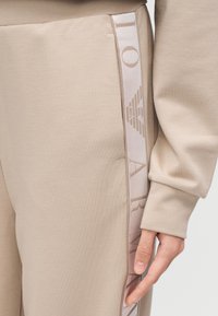 Μπεζ παντελόνια με ομαλή υφή διαθέτουν πλαϊνή λωρίδα με το λογότυπο «ARMANI» σε διακριτικό, ανοιχτόχρωμο τύπωμα.