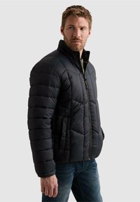 Homme regardant sur le côté portant une veste matelassée noire zippée avec des poches et un jean bleu sur un fond uni.