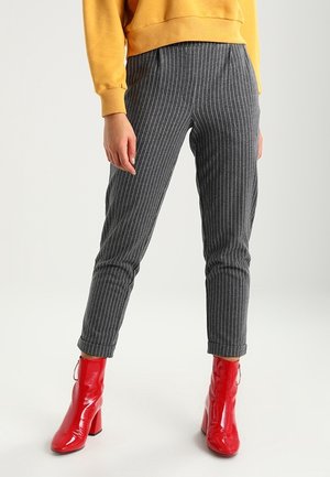 Kobieta ubrana w szare wąskie spodnie w prążki, musztardowożółtą bluzę oraz błyszczące czerwone botki na szerokim obcasie.