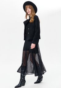 Zwarte getextureerde jas met een getailleerde pasvorm, over een doorschijnende zwarte kanten rok gedragen. Gecombineerd met zwarte kniehoge laarzen met hak en een hoed met brede rand.