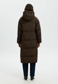 Abrigo de plumón marrón con capucha, con un diseño acolchado y un corte a la altura de la rodilla. Tejido texturizado y mangas largas. Jeans negros y botas de tobillo visibles.