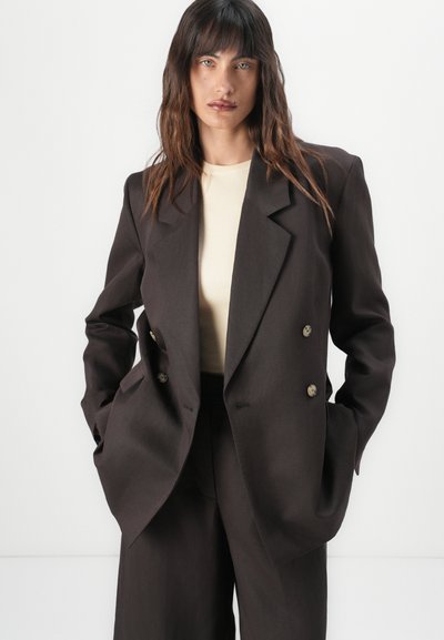 Blazer doppiopetto marrone scuro con ampie rever, tasche frontali e bottoni dorati, abbinato a un top beige chiaro. Texture liscia.