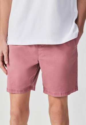 Mand iført rosafarvede shorts og en hvid skjorte, hænderne i lommerne, stående mod en ensfarvet lys baggrund.