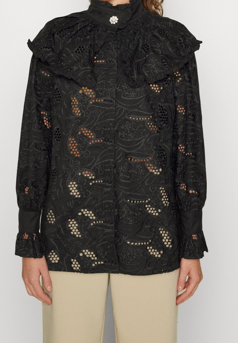 Blouse noire avec un col à volants, broderie florale délicate, détails en ajouré et manches longues avec poignets froncés.