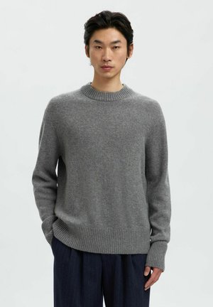 Jonge man in een grijze gebreide trui en donkere gestreepte broek, staand tegen een effen witte achtergrond.