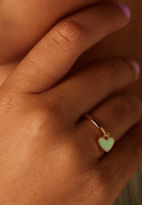 Goudkleurige ring met een mintgroene hartvormige emaille accent. De hand vertoont een natuurlijke textuur en heeft een neutrale nagellak kleur.