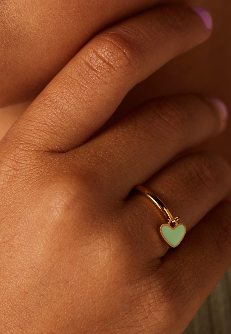 Goudkleurige ring met een mintgroene hartvormige emaille accent. De hand vertoont een natuurlijke textuur en heeft een neutrale nagellak kleur.