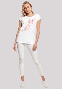 Weißes, kurzärmliges T-Shirt mit floralem Design in Rosa und japanischem Text. Kombiniert mit weißen Slim-Fit-Hosen und weißen Sneakern.
