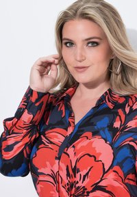 Kauluspaita, jossa on rohkea kukkakuosi punaisena, sinisenä ja mustana, sileä kankaan rakenne ja nappiedustukset hihoissa.