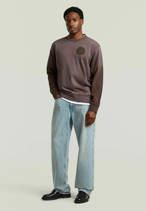 Homme debout, main gauche dans la poche, portant un sweat-shirt marron foncé, un jean large bleu clair et des chaussures noires sur un fond uni.