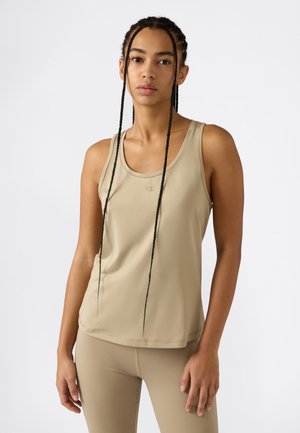 Jonge vrouw met lange vlechten, gekleed in een beige mouwloze top en bijpassende legging, staand tegen een effen witte achtergrond.