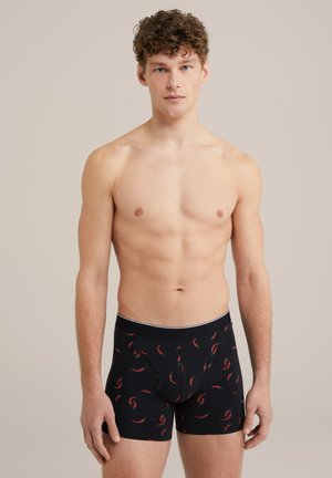 Fekete boxer alsónadrág, chili paprika mintával, puha anyagból készült, kontrasztos derékpánttal. A modellen látható, hogy ing nélkül áll.
