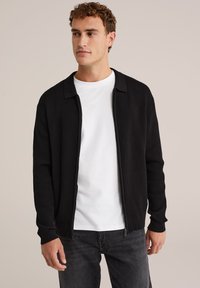Zwarte geribde cardigan met een kraaghals en volledige rits. Draagt over een wit t-shirt, gecombineerd met een donkere spijkerbroek. Speciale gladde textuur.