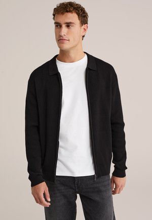 Cárdigan negro de canalé con cuello y cremallera completa. Llevado sobre una camiseta blanca, combinado con jeans oscuros. Textura suave.