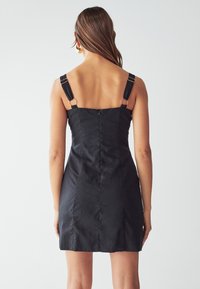 Musta mekko, jossa on vartalonmyötäinen muotoilu, säädettävät hihnat metallikoristeilla ja vetskari takana. Silkkinen, teksturoitu kangas.