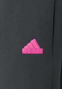 Zwarte sportbroek met een getextureerd roze Adidas-logo. Het ontwerp bevat een klassieke accent van drie strepen nabij het logo.