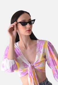 Occhiali da sole neri a forma di occhio di gatto con montatura angolare. Il modello indossa una camicetta bianca con un motivo tie-dye rosa e giallo, annodata sul davanti.