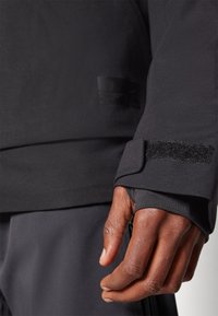 Black athletic jacket with a smooth texture, Velcro wrist cuffs, and a subtle logo detail. The fabric appears lightweight and flexible.