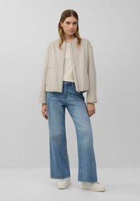 Giacca bomber beige con zip frontale, tasche laterali e una texture morbida, abbinata a jeans a gamba larga in un lavaggio chiaro e sneakers bianche.