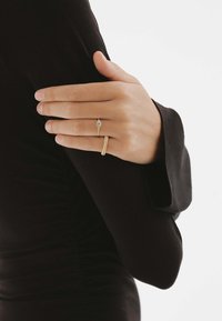 Anillo de oro con una pequeña gema azul y una banda texturizada, llevado en una mano apoyada sobre un fondo de tela negra.