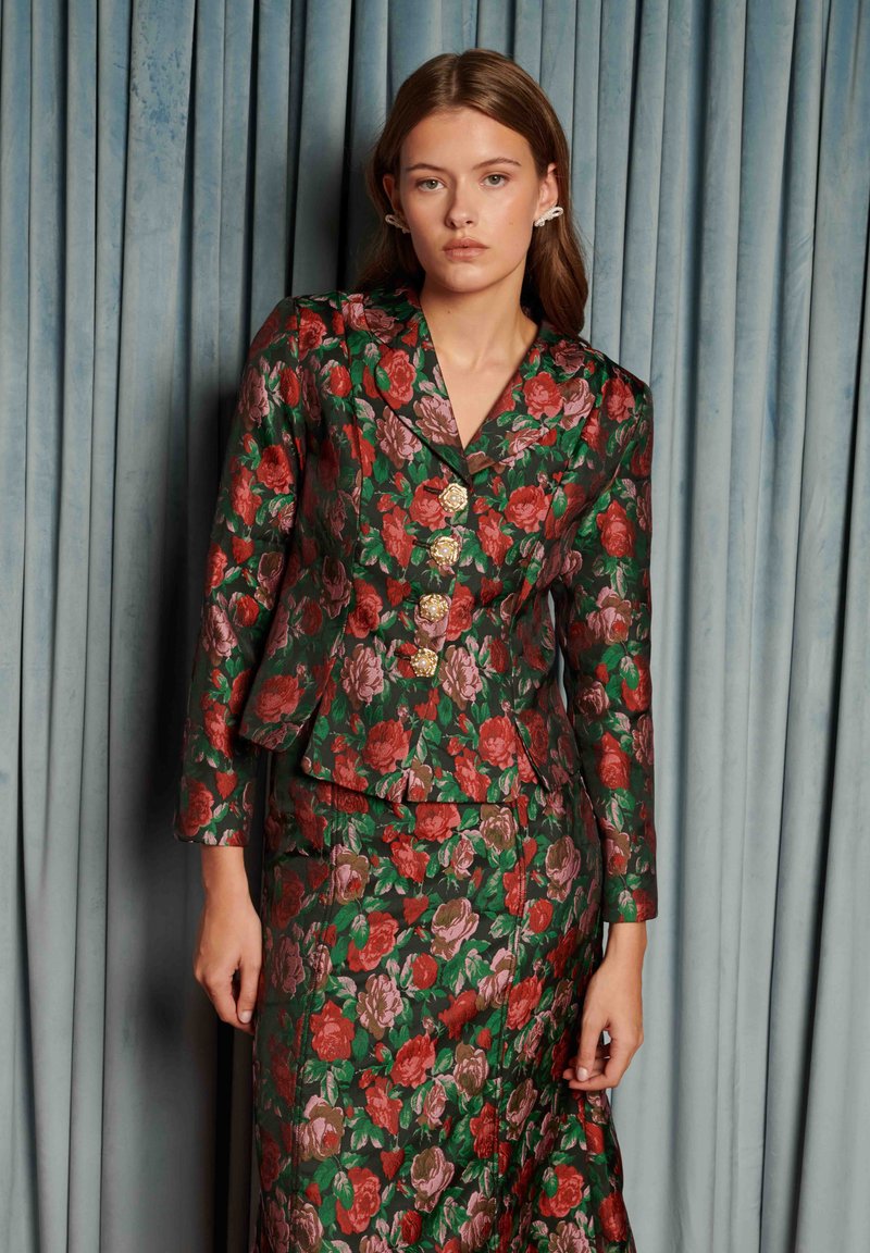 Lillemustriga ülikond läikivast kangast; tumeroheline punaste ja roosade roosidega; sisaldab liibuvat bleiserit kuldsete roosikujuliste nööpidega ja sobivat seelikut.