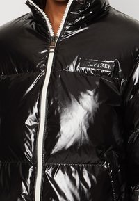 Sort, blank pufferjakke med høj krave, hvid lynlås og logo-patch på brystet. Har quiltet tekstur og vandrette syninger.