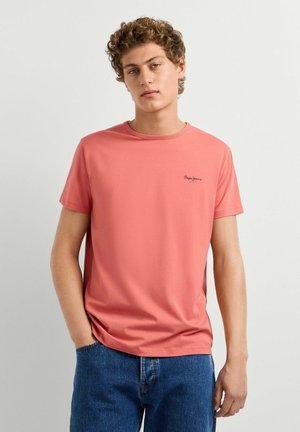 Giovane uomo con capelli ricci che indossa una maglietta rosa salmone a maniche corte e jeans blu, in piedi davanti a uno sfondo chiaro e uniforme.