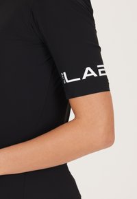 Svart träningsöverdel med korta ärmar, med vit text "ELITE LAB" på ärmen. Slät, elastisk tyg med en åtsittande design.