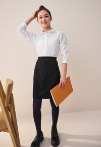 Hvid skjorte, sort nederdel og sorte strømpebukser. Kvinde holder en orange mappe. Har et enkelt, skræddersyet design. Træstol i baggrunden.