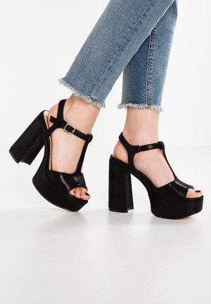 Černé platformové sandály na vysokém podpatku mají design s T-páskem, texturovaný materiál a zapínání na přezku. Otevřená špička a masivní blokové podpatky.