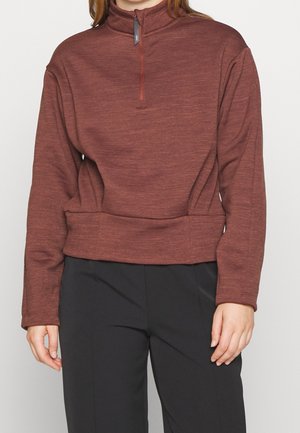 Personne portant un pull bordeaux à manches longues avec une fermeture éclair au col et un pantalon noir, debout sur un fond uni.