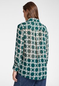 Camicia a maniche corte in tessuto verde trasparente con motivi geometrici bianchi. Presenta un colletto, una pattina con bottoni e una vestibilità leggermente comoda.