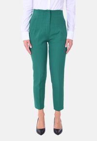 Pantaloni verdi a vita alta con pieghe, gambe affusolate e una texture liscia, abbinati a tacchi neri e una camicia bianca con bottoni.