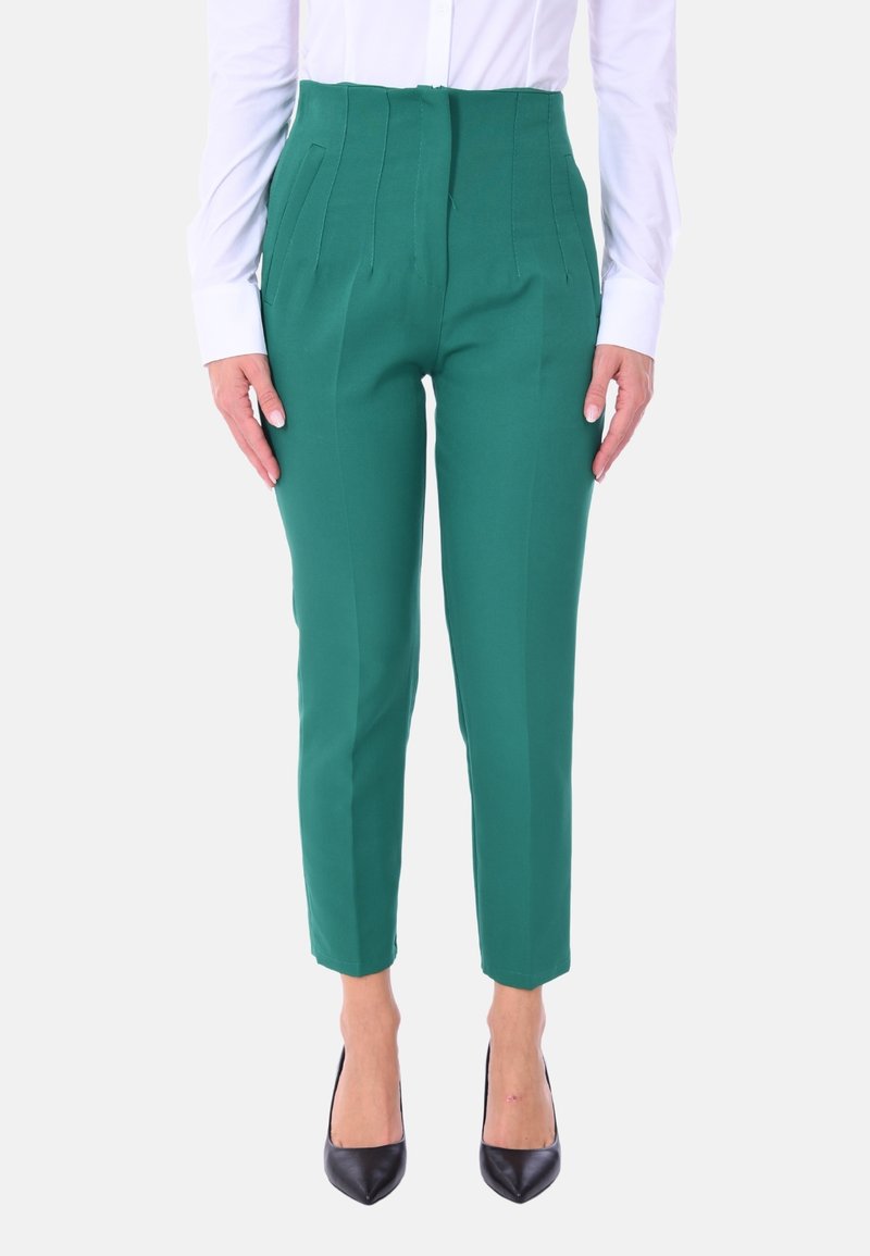 Pantaloni verdi a vita alta con pieghe, gambe affusolate e una texture liscia, abbinati a tacchi neri e una camicia bianca con bottoni.