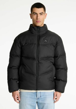 Hombre vestido con una chaqueta negra acolchada con cremallera frontal y jeans de color azul claro, de pie frente a un fondo blanco.