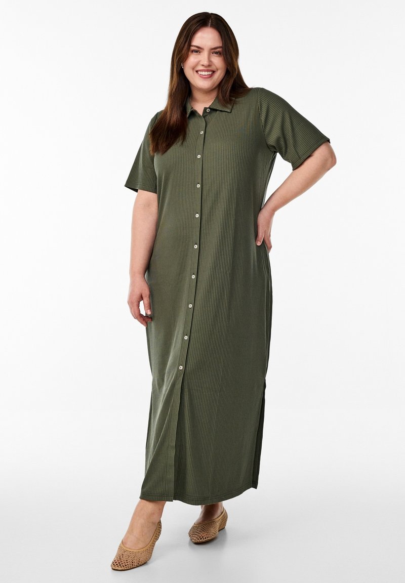 Vrouw lacht en staat met haar hand op haar heup, draagt een lange olijfgroene jurk met knopen en beige geweven platte schoenen.