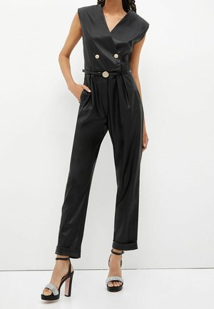 Černý bezramenní overal s překládaným designem, dvojitými knoflíky a páskem v oblasti pasu. Má hladký, lesklý materiál. V kombinaci s černými podpatky.