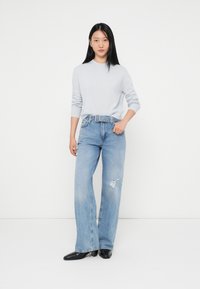Ljust grå tröja med ribbade manschetter, matchad med ljusblå vida jeans med slitningar. Modellen bär svarta skor.