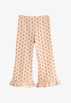 Ung pige med langt mørkt hår smiler iført en gul t-shirt med ananas og mønstrede vidde bukser med flæsekant og sandaler.