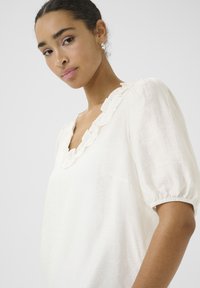 Blouse blanche avec un col en V ondulé et des manches courtes bouffantes. Fabriquée en tissu léger, avec une surface douce et texturée.