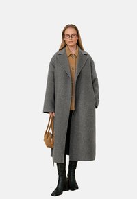 Cappotto di lana grigio con collo a sciallata, taglio oversize e bottoni frontali. Abbinato a una camicia marrone, pantaloni neri e stivali neri alti fino al ginocchio.