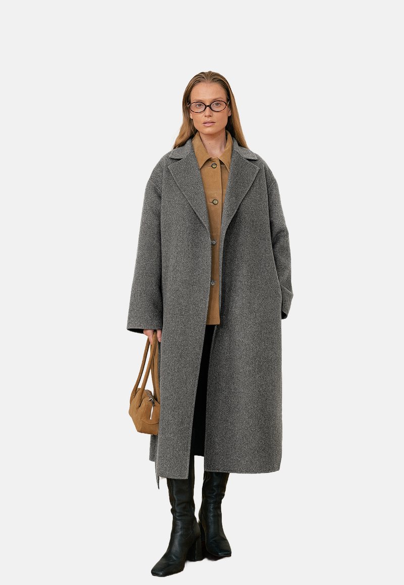 Cappotto di lana grigio con collo a sciallata, taglio oversize e bottoni frontali. Abbinato a una camicia marrone, pantaloni neri e stivali neri alti fino al ginocchio.