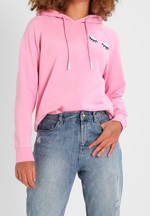 Kobieta w różowej bluzie z kapturem ze sznureczkami i grafiką rzęs na piersi, w połączeniu z jasnoniebieskimi, zniszczonymi jeansami z wysokim stanem.