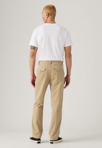 Beige katoenen broek met achterzakken en een klein logo-label, gecombineerd met een effen wit shirt en zwarte schoenen, gezien vanaf de achterkant.