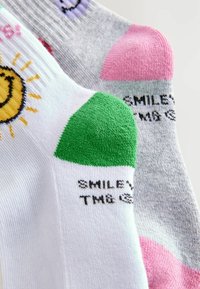Calze colorate con motivi: bianche con un sole sorridente, grigie con dettagli, tallone verde, punta rosa e testo nero "SMILEY." Tessuto testurizzato.