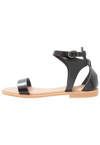 Mustat nahkasandaalit, joissa on tasapohja, leveä etuosa ja nilkkasanko metallisoljella. Minimaalinen muotoilu avoimilla sivuilla.